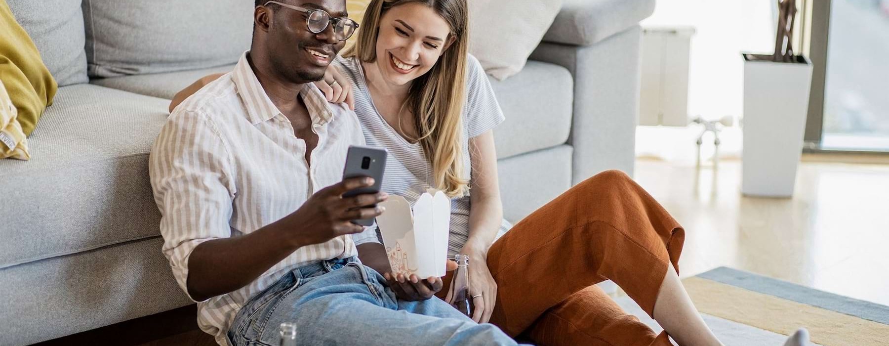 a man and woman with take out, sit against a couch on their living room floor and watch at their cell phone